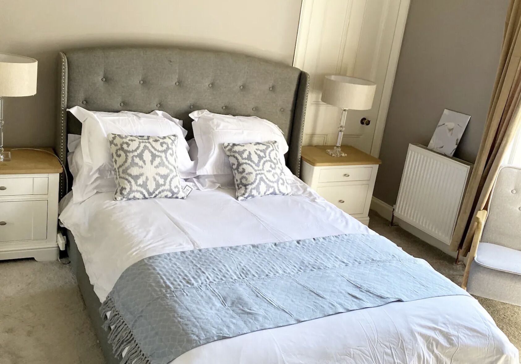 A neatly made bed with a grey upholstered headboard, white linens, and patterned pillows. There are two bedside tables with lamps, a radiator, and a door visible.