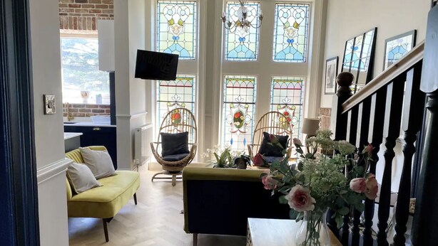 Ein gemütliches Wohnzimmer mit einem gelben Sofa, Korbstühlen, einem Holztisch und einem Blumenstrauß.