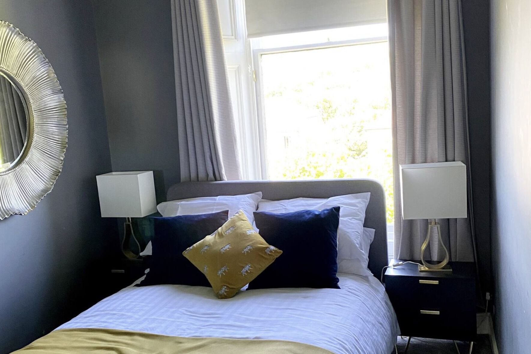 A neatly made bed with white and navy bedding, two bedside lamps, and a large window with curtains.