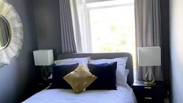 A neatly made bed with white and navy bedding, two bedside lamps, and a large window with curtains.