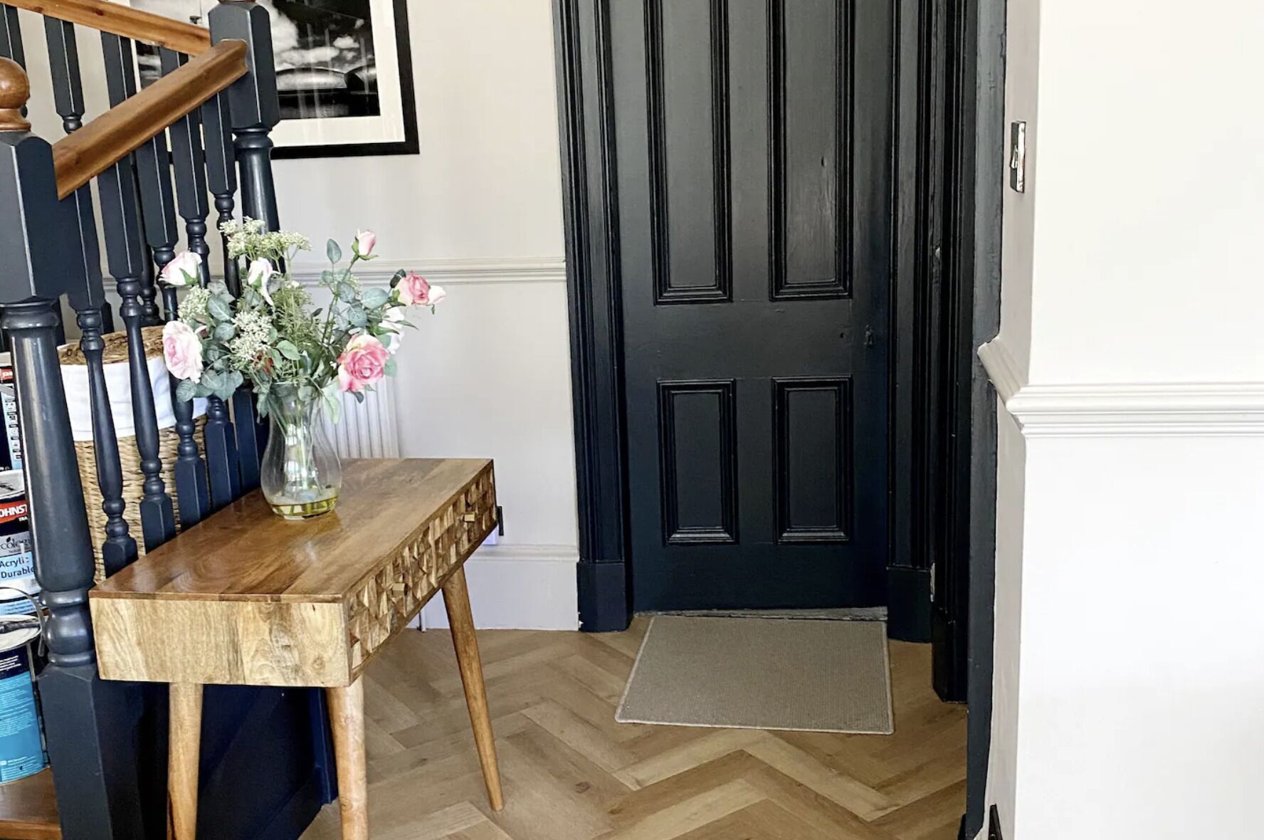 Una consola de madera con un jarrón de flores, una puerta negra y un suelo con patrón de espiga.