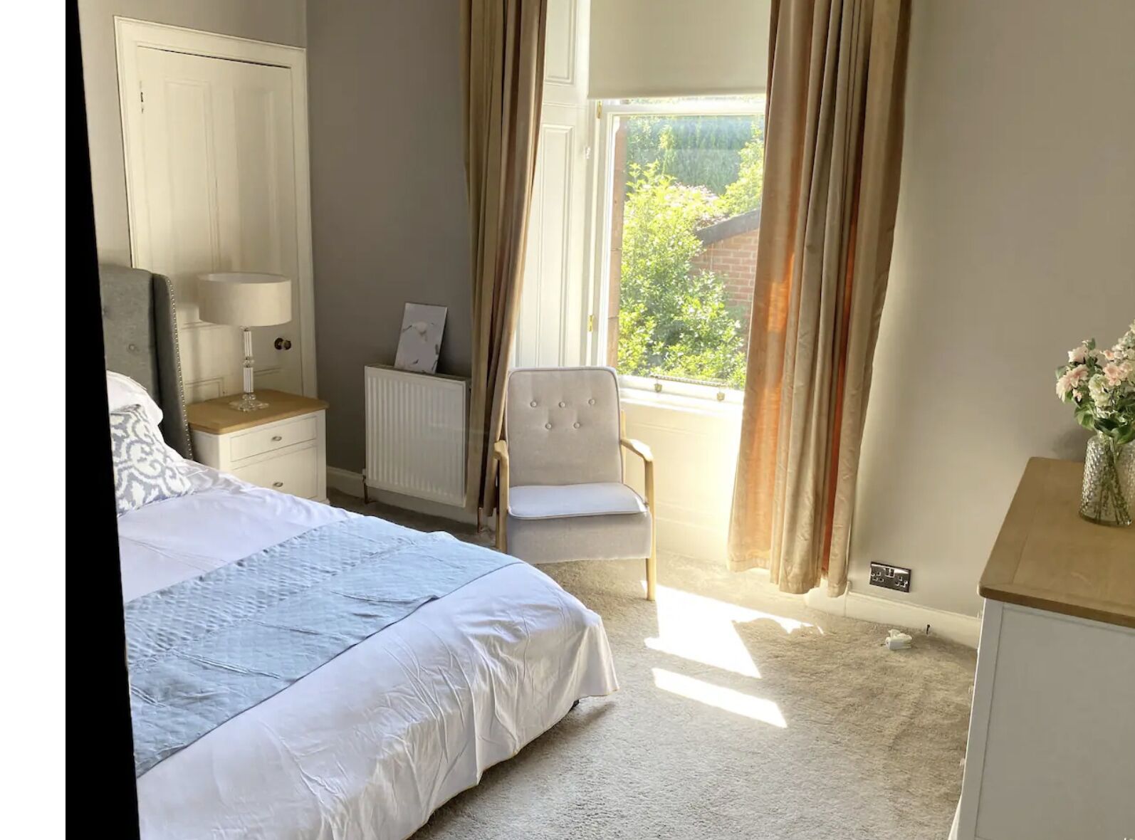 A bedroom with a bed, bedside table, chair, radiator, and a window with curtains.