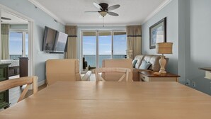 A room with a large window, a ceiling fan, a TV, a table, and two chairs. - Emerald Isle 2110 - Front row beach chair service included! Affordable rates (Panama City Beach)