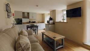 Ein Wohnzimmer mit einer Couch, einem Couchtisch, einem an der Wand befestigten Fernseher, einer Küchenzeile mit Schränken und Geräten sowie einem Esstisch mit Stühlen.