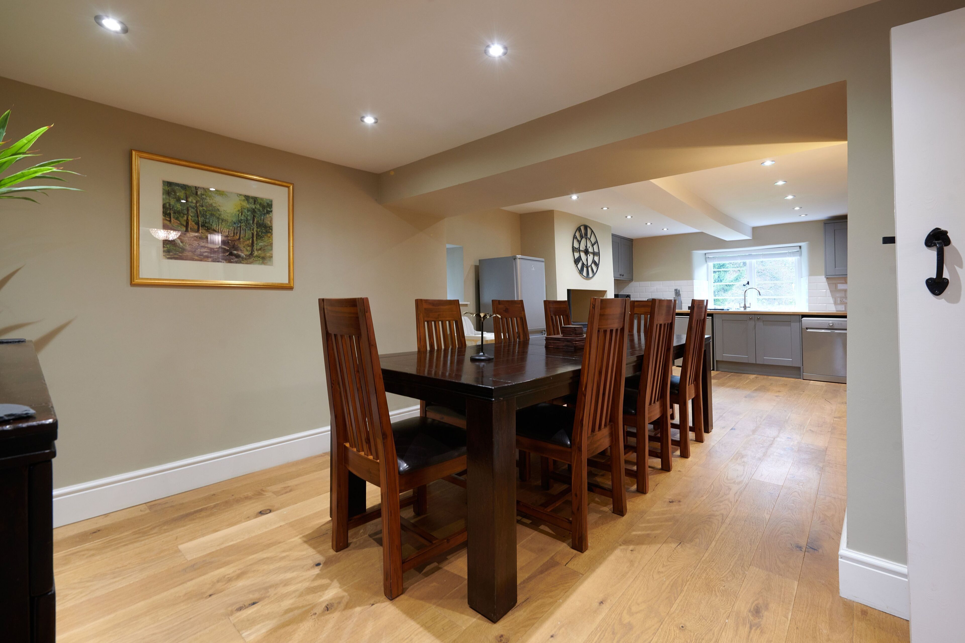 A dining area with a wooden table and chairs, a framed picture on the wall, and a kitchen in the background.