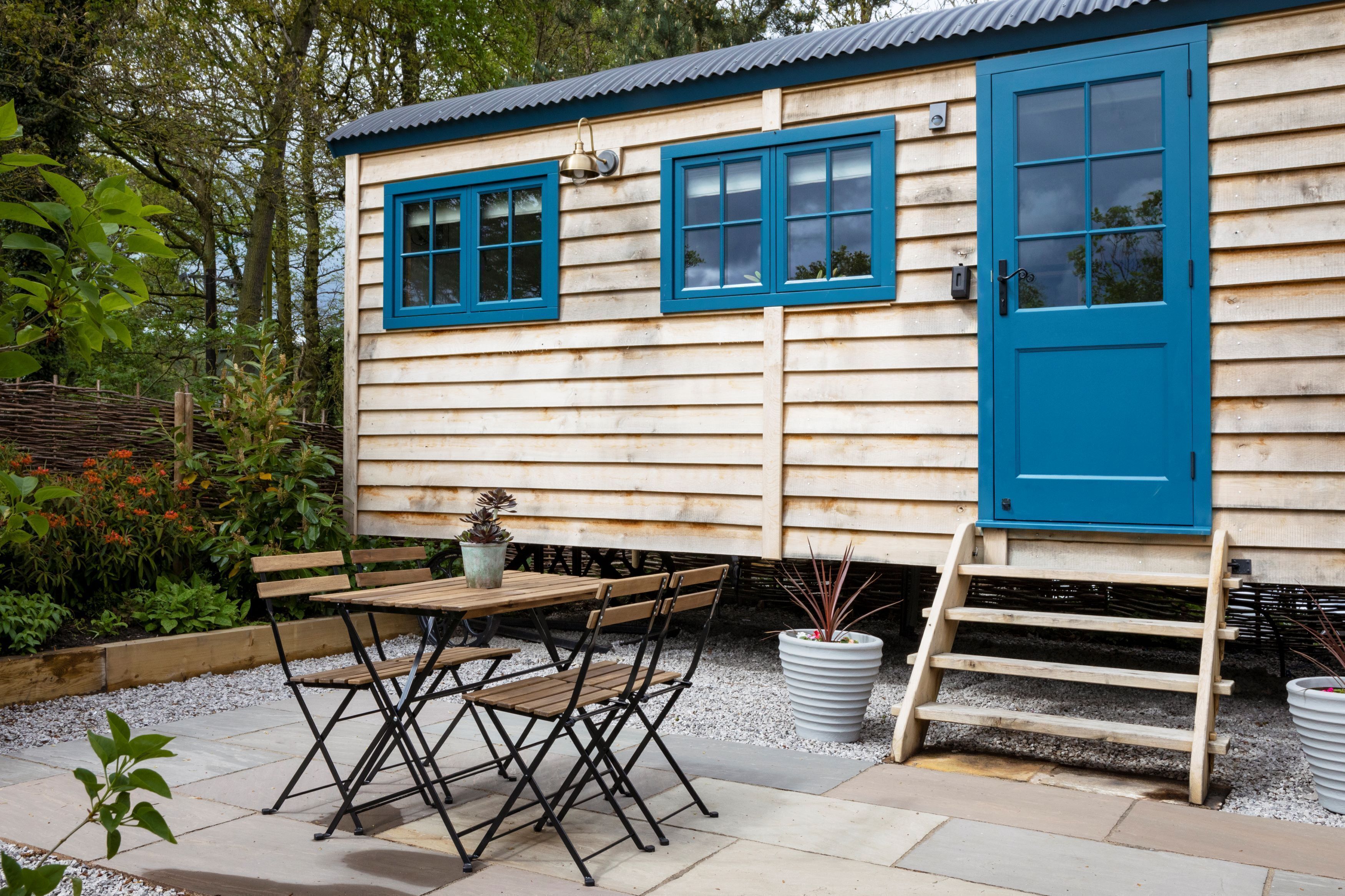 A wooden cabin with a blue door and windows, a wooden table with chairs, and a small garden area with plants and a potted plant.