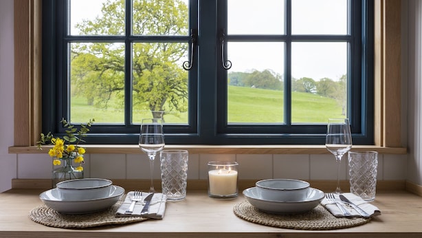 A dining area with a table set for two, a vase of yellow flowers, and a view of a green landscape through a window.
