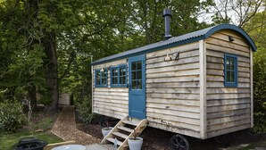 A small wooden cabin with a blue door and windows, a chimney, and a small patio with a table and chairs.