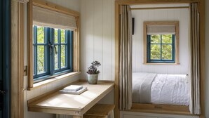 A room with a bed, a desk with a plant, and two windows with curtains. - Woodlands Shepherd's Hut Chatsworth Estate (Baslow)
