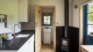 A kitchen with a black stove, white cabinets, a wooden door, and a window with a view. - Woodlands Shepherd's Hut Chatsworth Estate (Baslow)