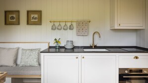 A kitchen with white cabinets, a black countertop, a sink, and a stove. There are two framed pictures on the wall, a vase with flowers, and a set of mugs hanging above the sink. - Woodlands Shepherd's Hut Chatsworth Estate (Baslow)
