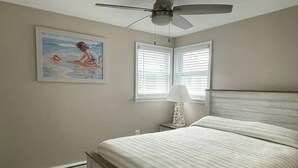A bedroom with a bed, a ceiling fan, a nightstand with a lamp, and a painting on the wall. - 231 33rd St Unit A  2BR Beach Home Hideaway  Parking (Brigantine)