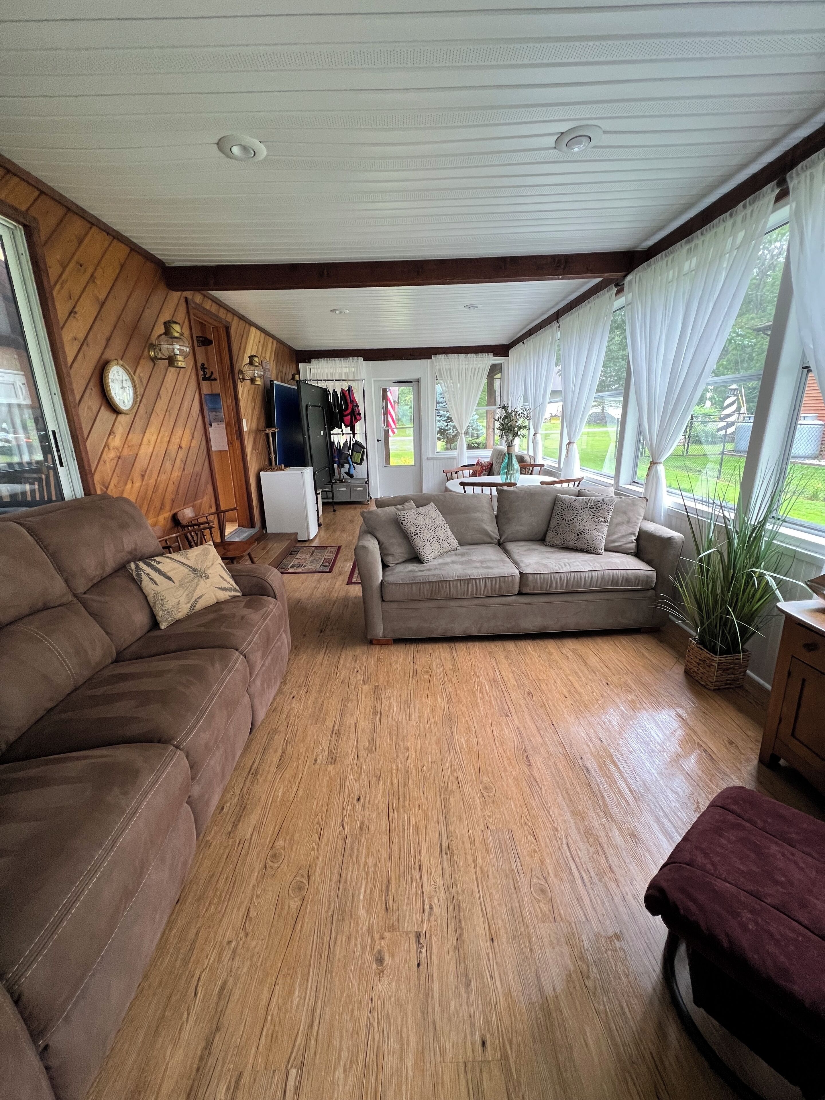 A cozy living room with a wooden floor, a brown sofa, a grey sofa, and a red ottoman. There is a dining table with chairs, a white ceiling fan, and a window with white curtains.