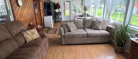 A cozy living room with a wooden floor, a brown sofa, a grey sofa, and a red ottoman. There is a dining table with chairs, a white ceiling fan, and a window with white curtains.