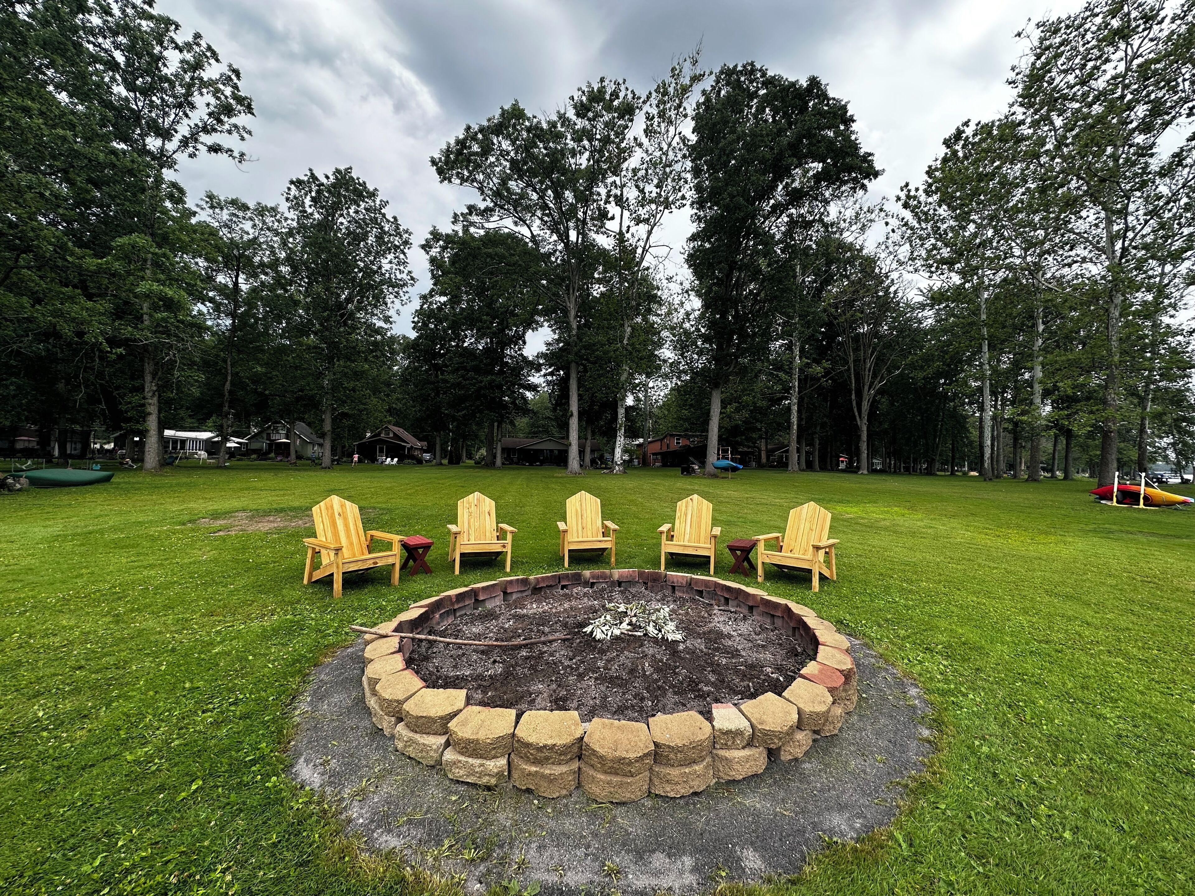 A fire pit surrounded by wooden chairs in a grassy yard with trees and houses in the background.