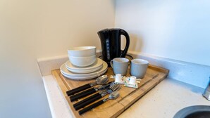 Eine Küchenarbeitsplatte mit einer Stapel weißer Teller, einer schwarzen Kaffeemaschine, grauen Tassen und Besteck auf einem Holztablett.