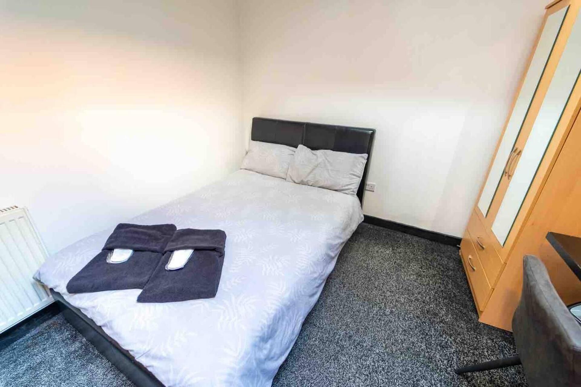 Un petit lit bien fait, avec une couette grise et deux oreillers, une table de chevet en bois avec une lampe, et un radiateur sur le côté gauche.