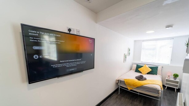 A modern hotel room with a bed, a bedside table, a small plant, and a TV mounted on the wall displaying a sign about Amazon account creation. - Large Studio Available on First Floor! (Birmingham)