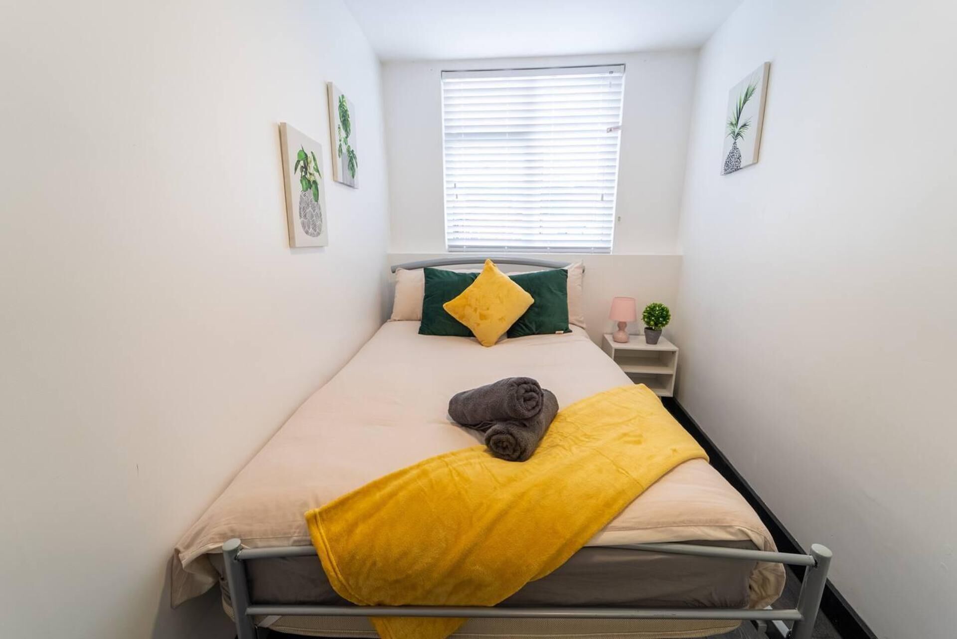 Une petite chambre avec un lit simple, une fenêtre, une table de chevet avec une lampe et des œuvres d’art accrochées au mur.