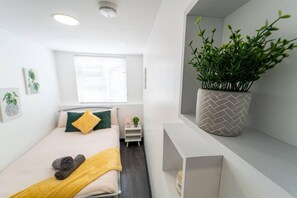 A modern, well-lit bedroom with a bed, a small bedside table, a potted plant, and a window with blinds. - Large Studio Available on First Floor! (Birmingham)