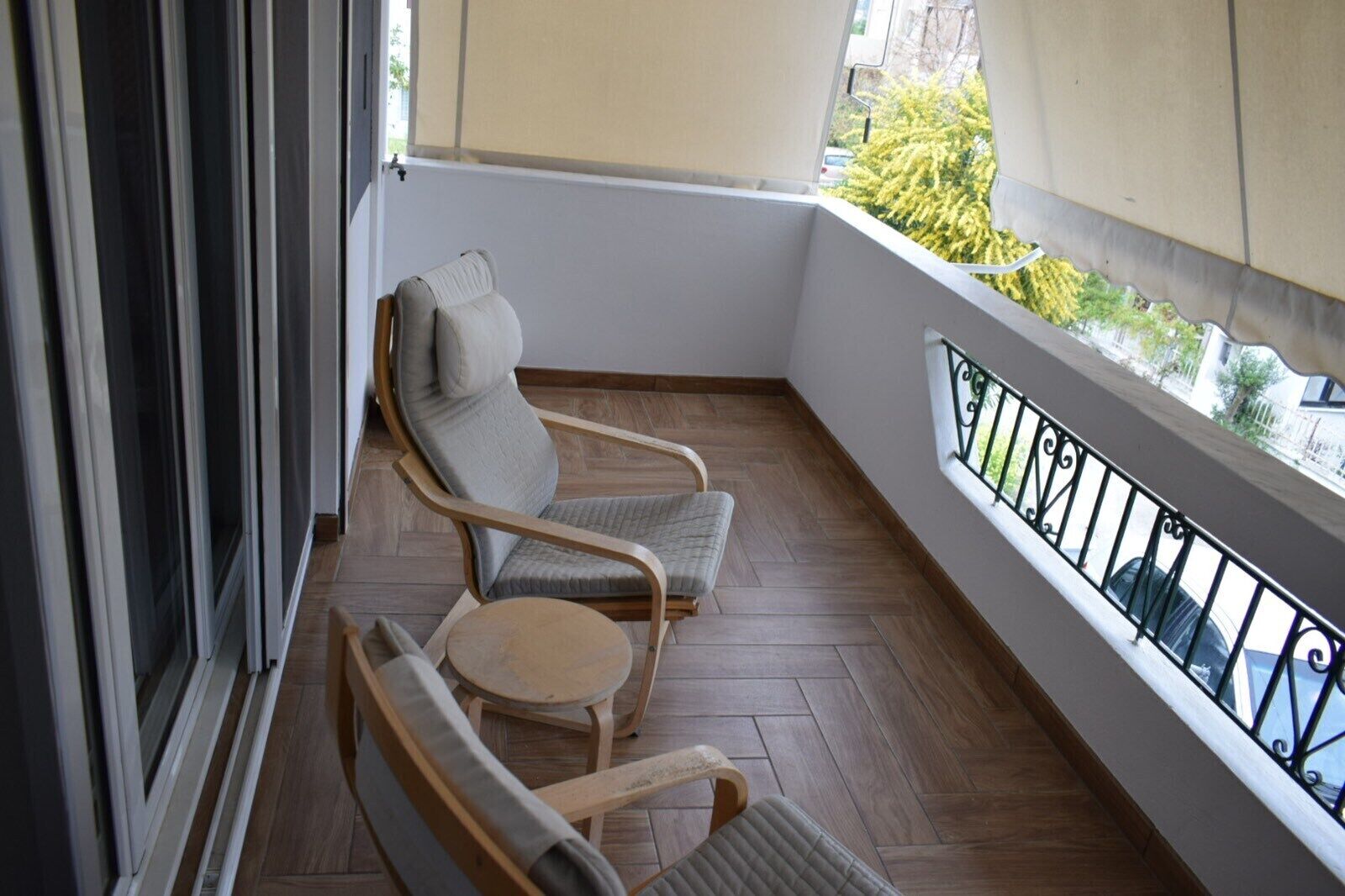 A balcony with two cushioned chairs and a small round table, wooden flooring, and a view of greenery outside.