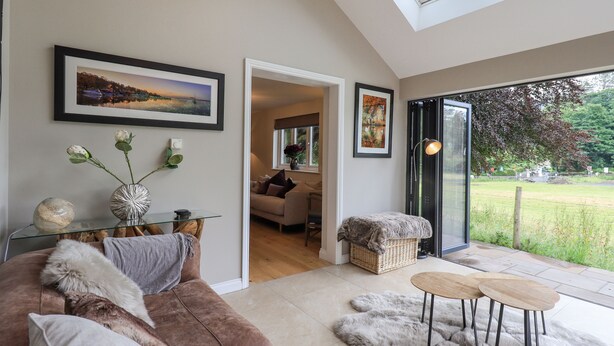 A modern living room with a brown sofa, a glass coffee table, and a fur rug. There is a framed painting on the wall and a window with a view of the outdoors. - Beech Lea (Ulverston)