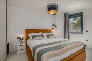 A modern bedroom with a wooden headboard, a large bed with striped bedding, a bedside table with a lamp, and a window with curtains. - Holiday apartment with pool (Njivice)