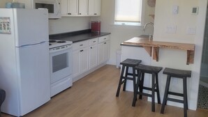 A compact kitchen with white cabinets, a white refrigerator, a stove, and a microwave. There are two wooden stools and a small window with blinds. - Cozy North Shore two bedroom cottage (York)