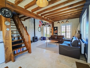 Uma sala de estar ampla com escada de madeira, lareira, sofás de couro e uma grande janela que oferece iluminação natural.