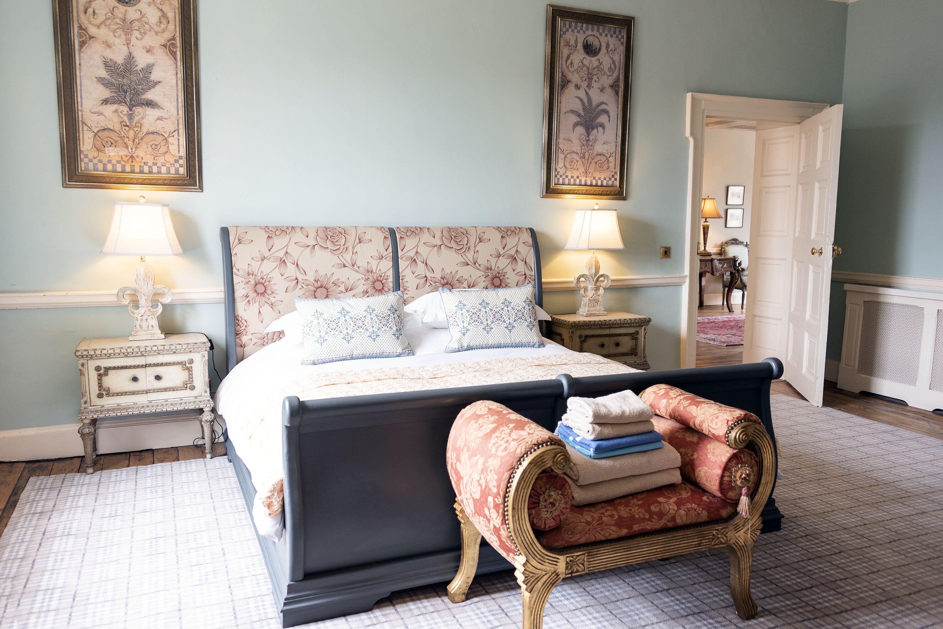 Un dormitorio con una cama grande, dos mesitas de noche con lámparas, una silla con toallas dobladas y una puerta que da a otra habitación.