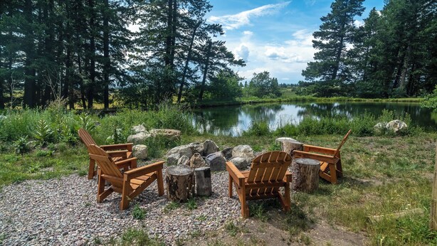 Eine Uferlandschaft mit Holz-Adirondack-Stühlen, die um eine Feuerstelle angeordnet sind, umgeben von Steinen und Bäumen.