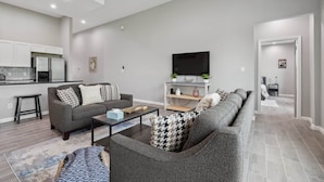 Ein modernes Wohnzimmer mit grauem Sofa, einem Couchtisch und einem Fernseher. Es gibt eine Küchenzeile mit weißen Schränken und einem Kühlschrank.