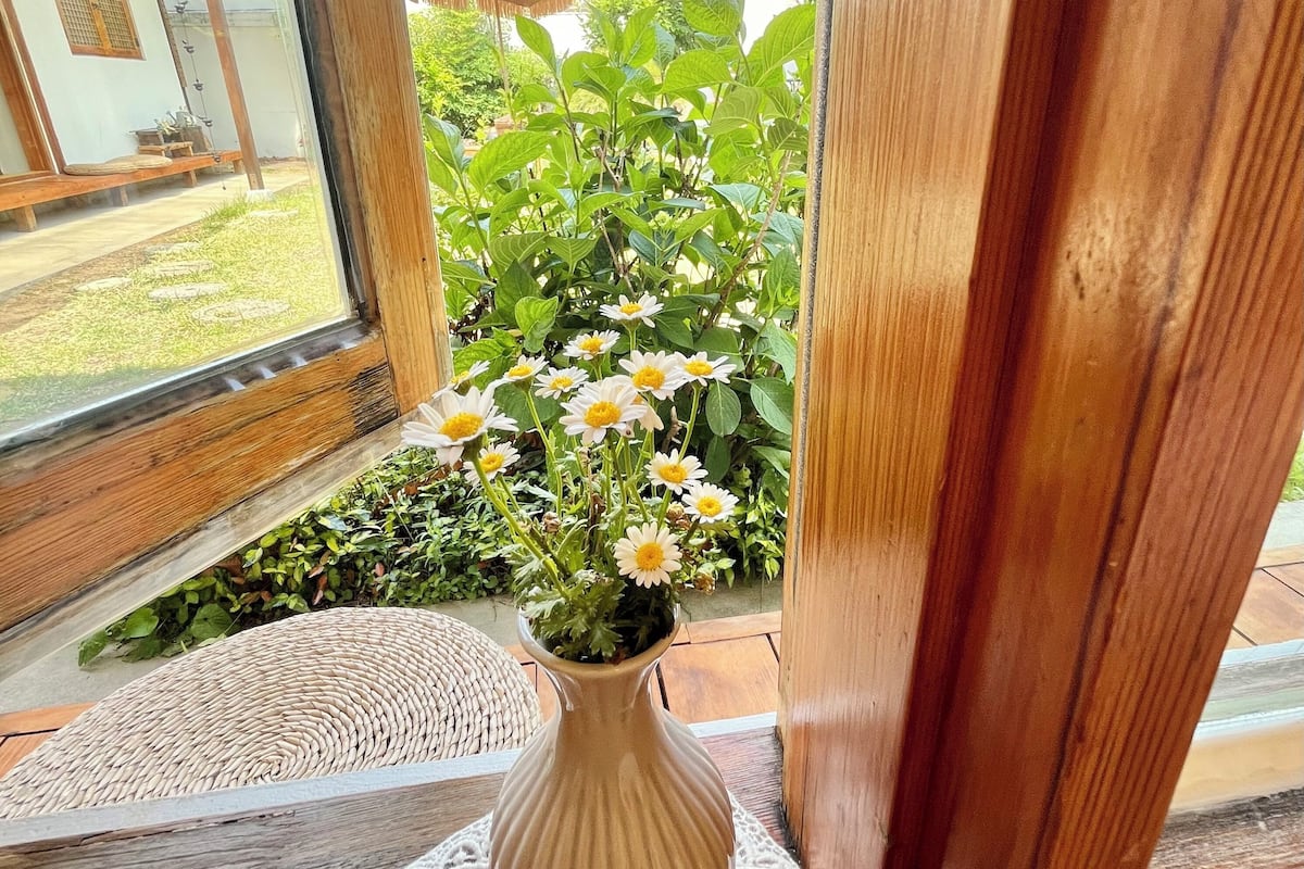 Ein Holsterfenster mit Blick auf einen Garten und eine weiße Vase mit Blumen auf einer Fußmatte.