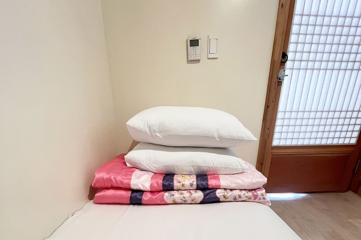 Ein kleines, ordentlich bezogenes Bett mit einer weißen Matratze und rosa Bettwäsche. Zwei Kissen, eines weiß und eines mit Blumenmuster. Der Raum hat eine Holztür mit einem Gittermuster und ein kleines, an der Wand befestigtes Gerät darüber.