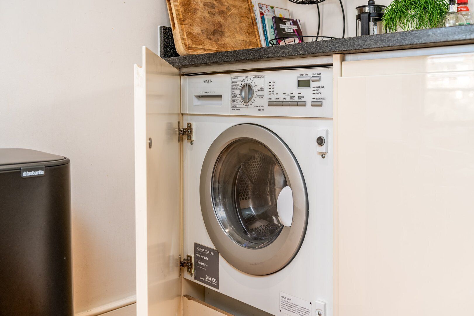 A front-loading washing machine in a compact kitchen area with a trash bin and a countertop with various items.