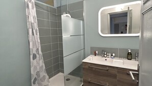 A modern bathroom with a glass-enclosed shower, a vanity with a mirror, and dark tiled flooring. - Très bel Appartement Neuf T4 (Toulon)