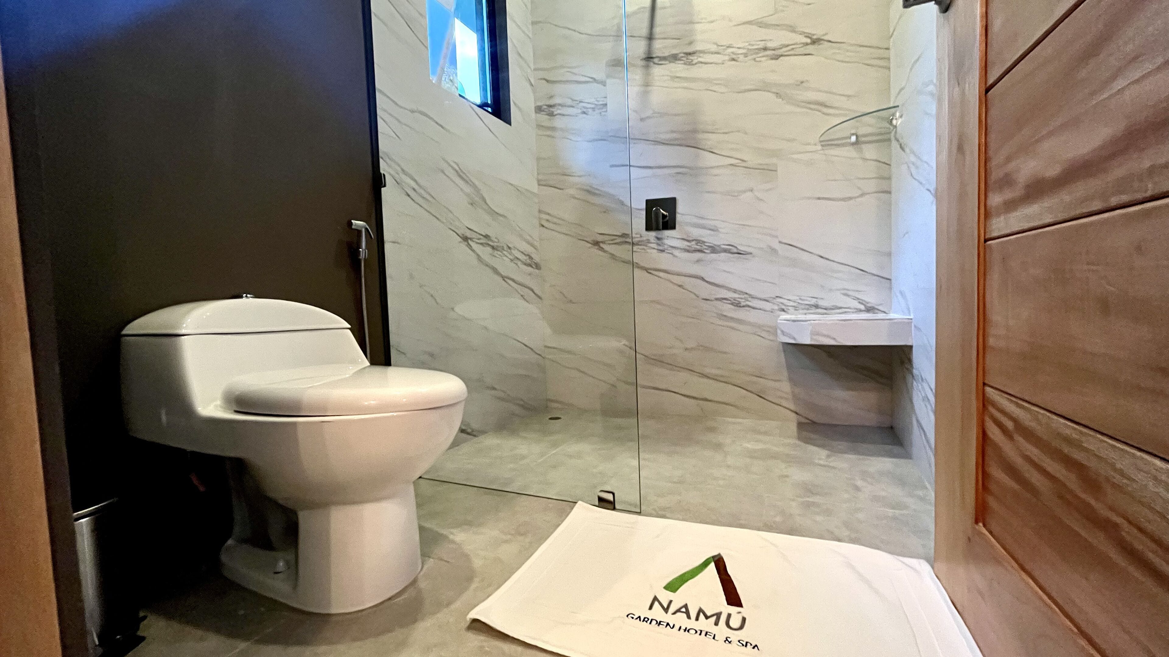 A modern bathroom with a toilet, a shower, and a sink. A white mat with a logo is on the floor.