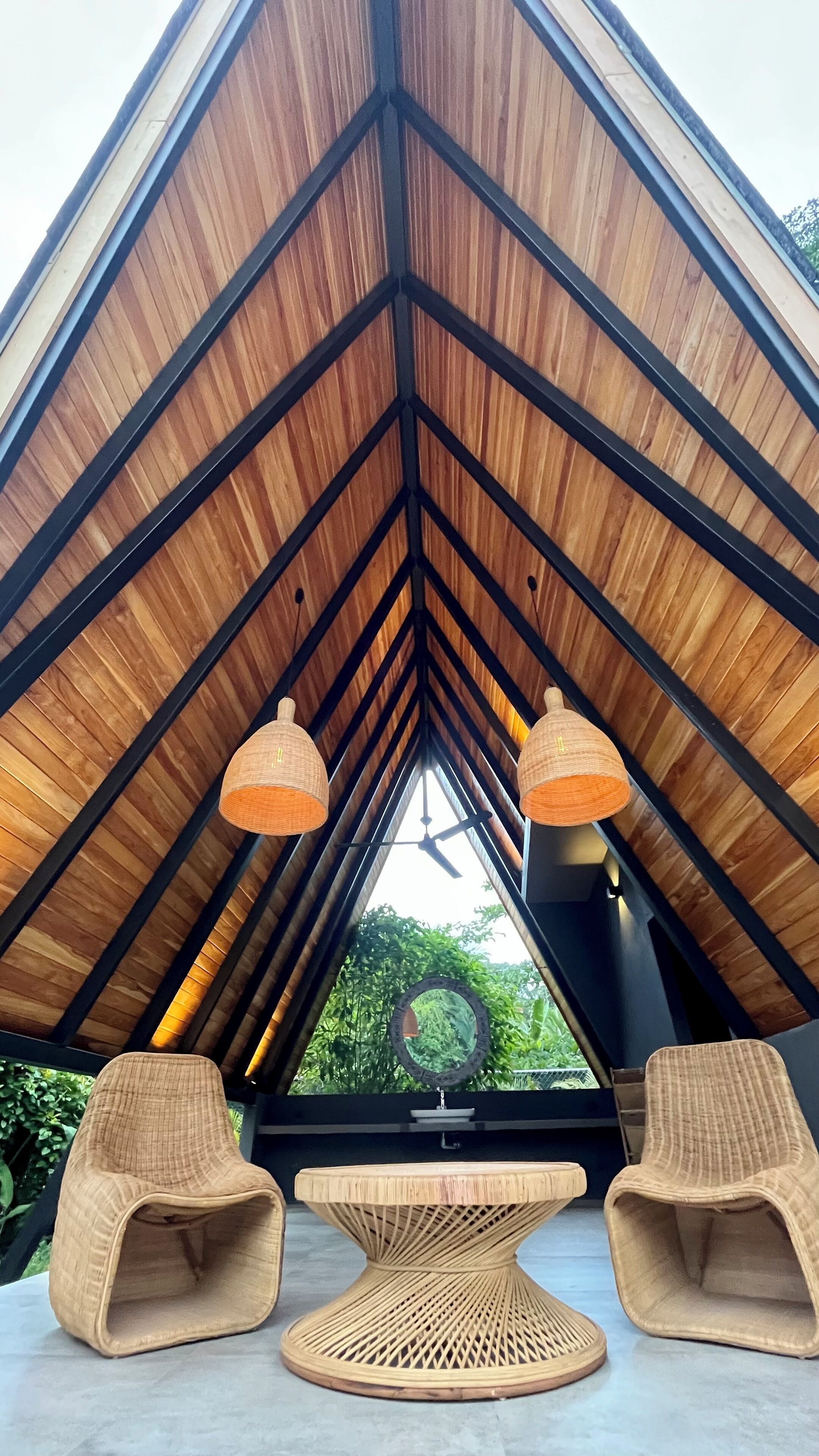 Une pièce dotée d’un plafond en bois incliné et très haut, de deux fauteuils en osier, d’une table centrale en fibres tressées et offrant une vue sur la verdure environnante.