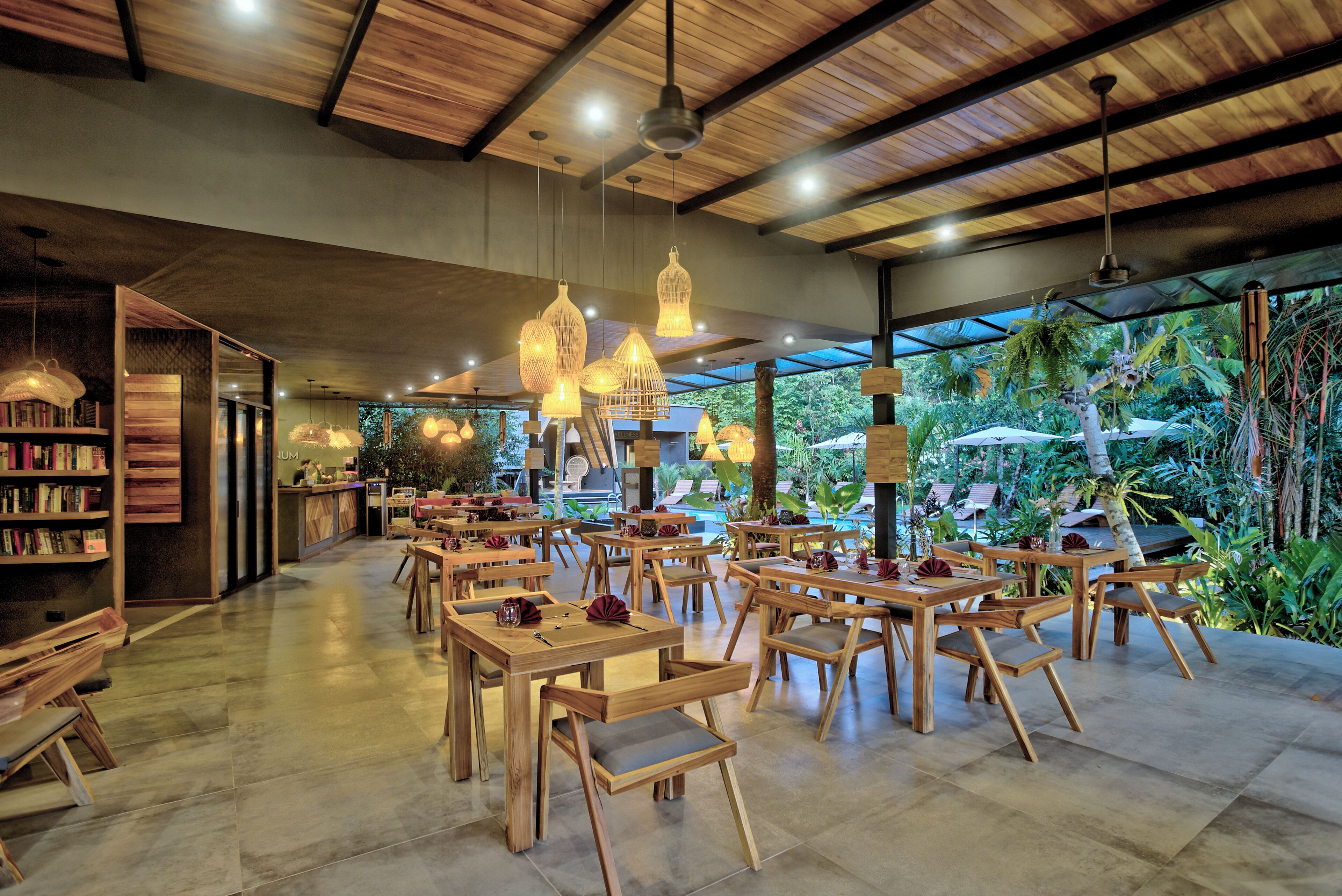 Un restaurante con mesas y sillas de madera, lámparas colgantes y vistas a zonas verdes.