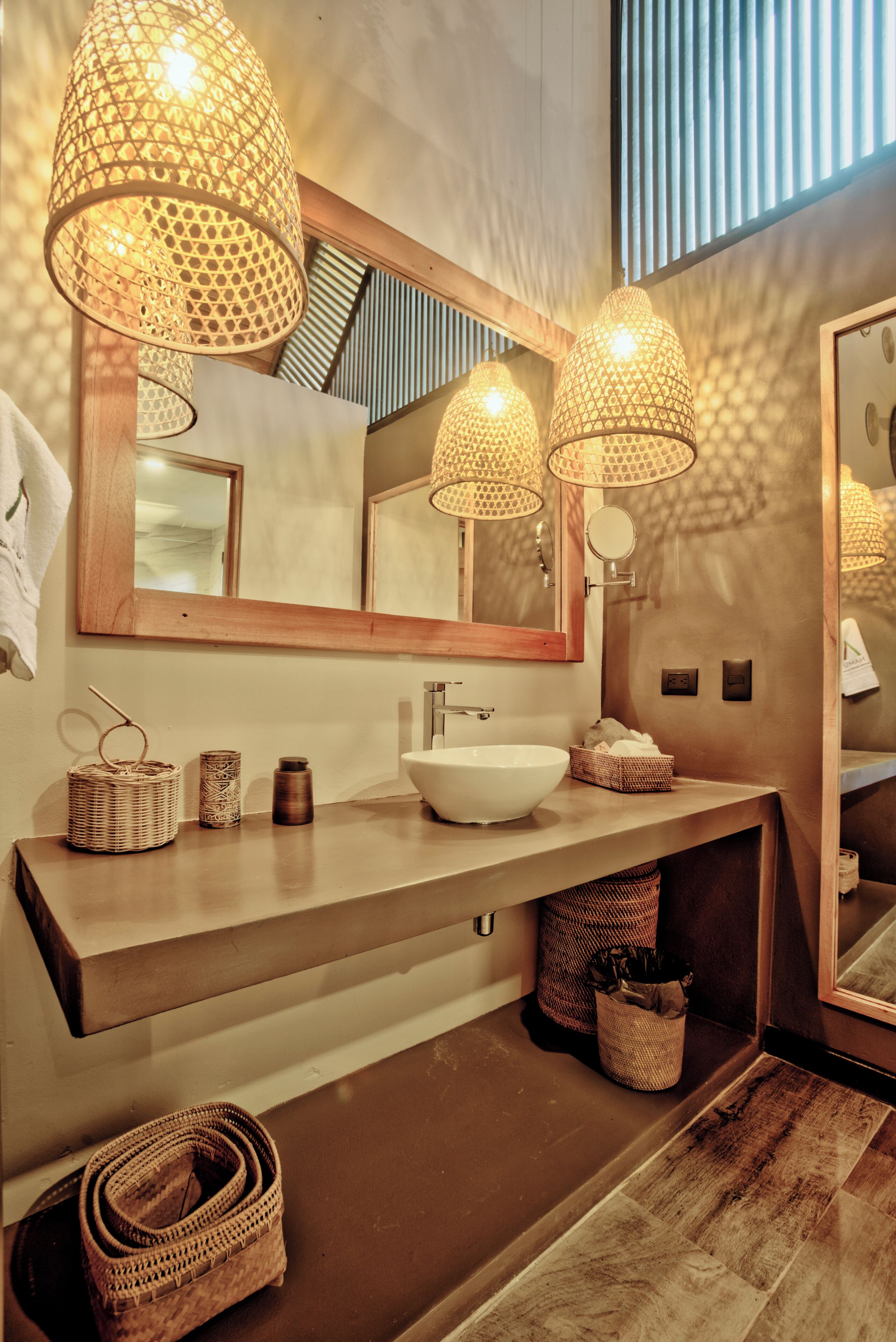 Une salle de bain moderne avec un grand miroir, un lavabo blanc et trois lampes suspendues. On y trouve des paniers en osier et un porte-serviettes sur le plan de travail.