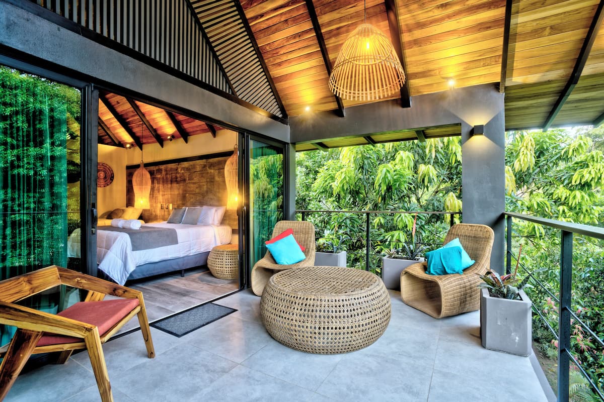 Une chambre moderne avec un mur en verre, un grand lit et un balcon agrémenté de meubles en osier et de plantes.