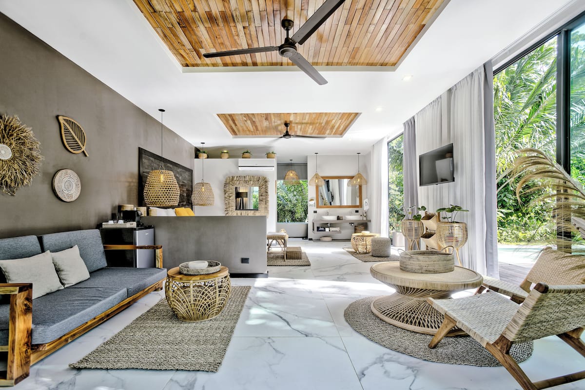 Un salon moderne avec un canapé, des meubles en osier, un ventilateur de plafond et une grande fenêtre donnant sur un paysage verdoyant.