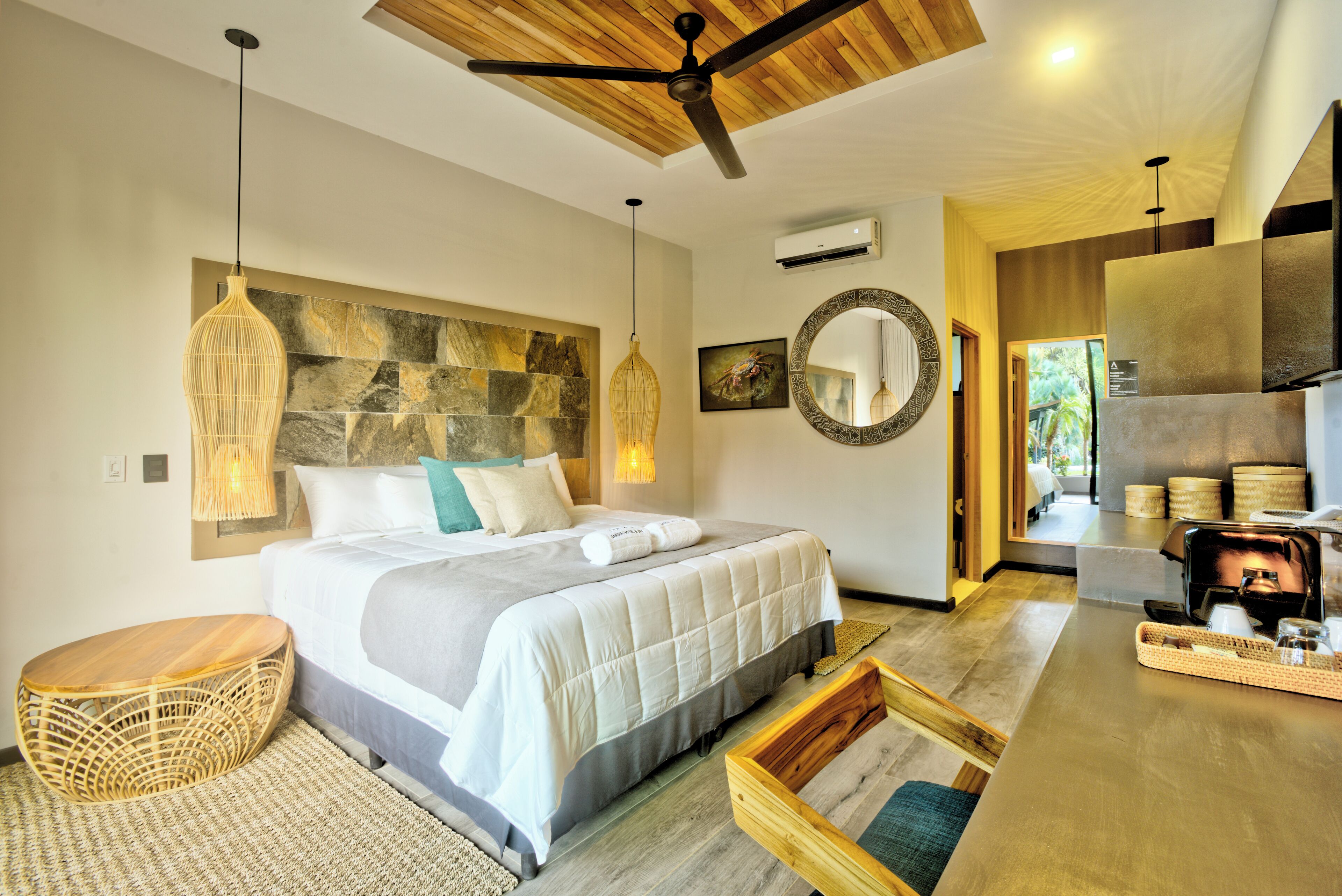 Une chambre moderne avec un grand lit, un ventilateur de plafond en bois, un mur d’accent en pierre, un miroir rond et une vue sur le salon.