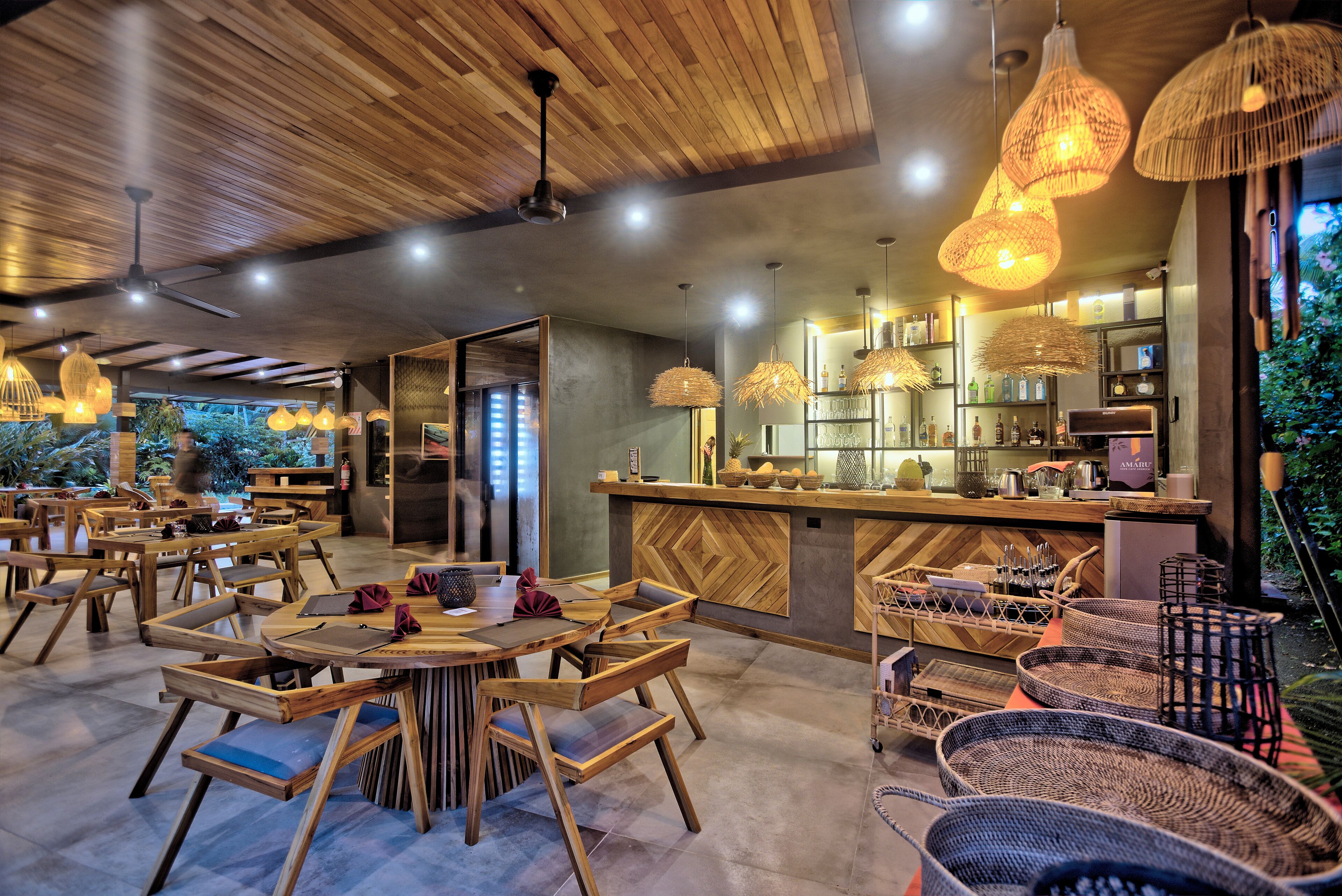 Un restaurante con muebles de madera, lámparas de mimbre colgantes y una zona de bar con estantes llenos de botellas.
