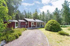 A red cabin with a porch and a gravel driveway, surrounded by greenery and trees.