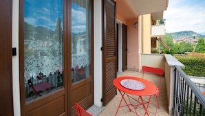 A balcony with a red table and chairs, a lace window treatment, and a view of a town and mountains in the background. - Appartamento La Rosa (Roe Volciano)