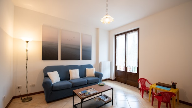 A living room with a blue sofa, a coffee table with magazines, a floor lamp, and a window with a view. - Appartamento La Rosa (Roe Volciano)