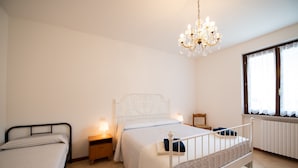 A bedroom with two beds, a chandelier, a window with lace curtains, and a bedside table with a lamp. - Appartamento La Rosa (Roe Volciano)