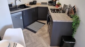 Eine kleine Küche mit dunklen Schränken, einer schwarzen Arbeitsplatte und einem kleinen Spülbecken. Auf der Arbeitsplatte befinden sich eine Kaffeemaschine, ein Toaster und ein Wasserkocher. Ein kleiner Teppich liegt auf dem Boden, und eine Pflanze steht auf der Arbeitsplatte.