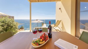 A table with a bottle of wine, two glasses of red wine, a plate of food, and a menu on a balcony overlooking the sea.